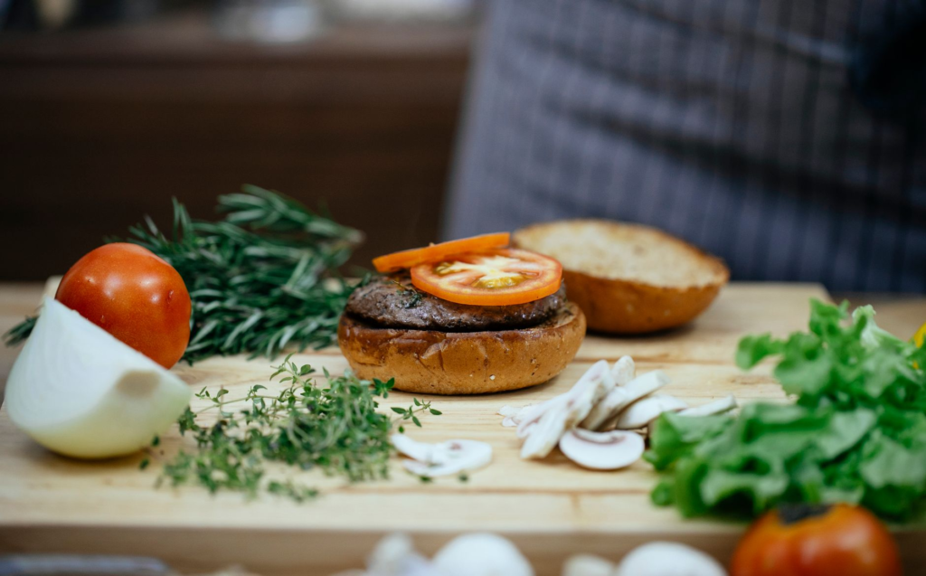 An assortment of traditional burger toppings like lettuce, tomatoes, and pickles displayed on a wooden cutting board. — …